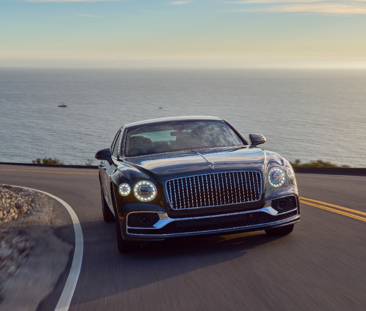 Black 2022 Bentley Flying Spur Hybrid driving a curved road with ocean in the background, front view