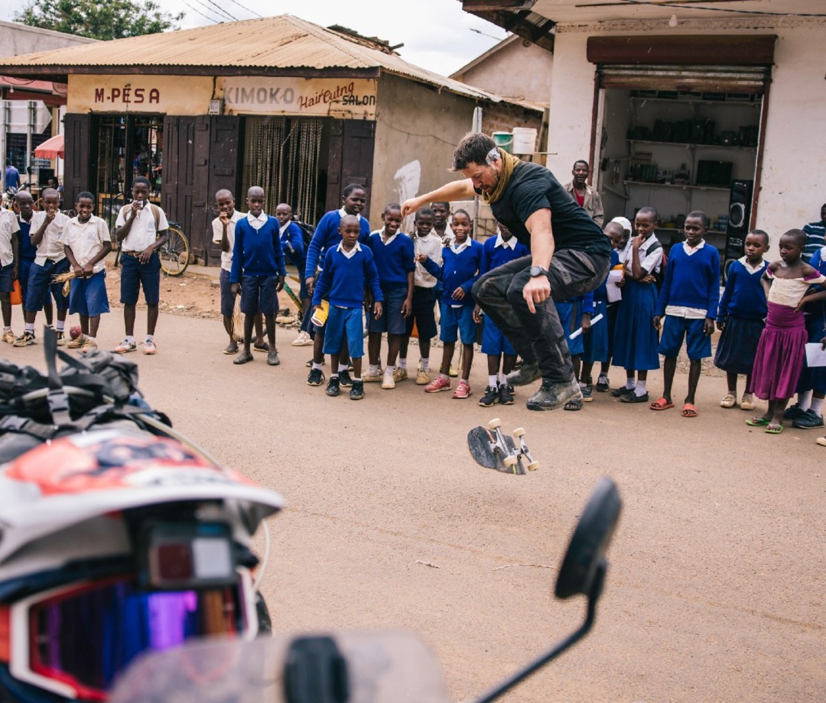 Motorcyclist performs skateboarding tricks for a crowd of young school kids in an African village