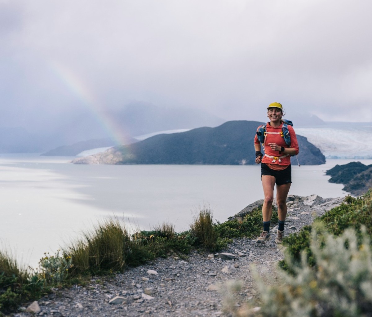 Woman jogs along a lakeside trail with rainbow in the background in Torres del Paine National Park