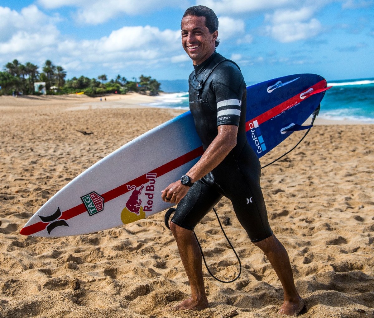A smiling Kai Lenny walks along the beach holding his surfboard.