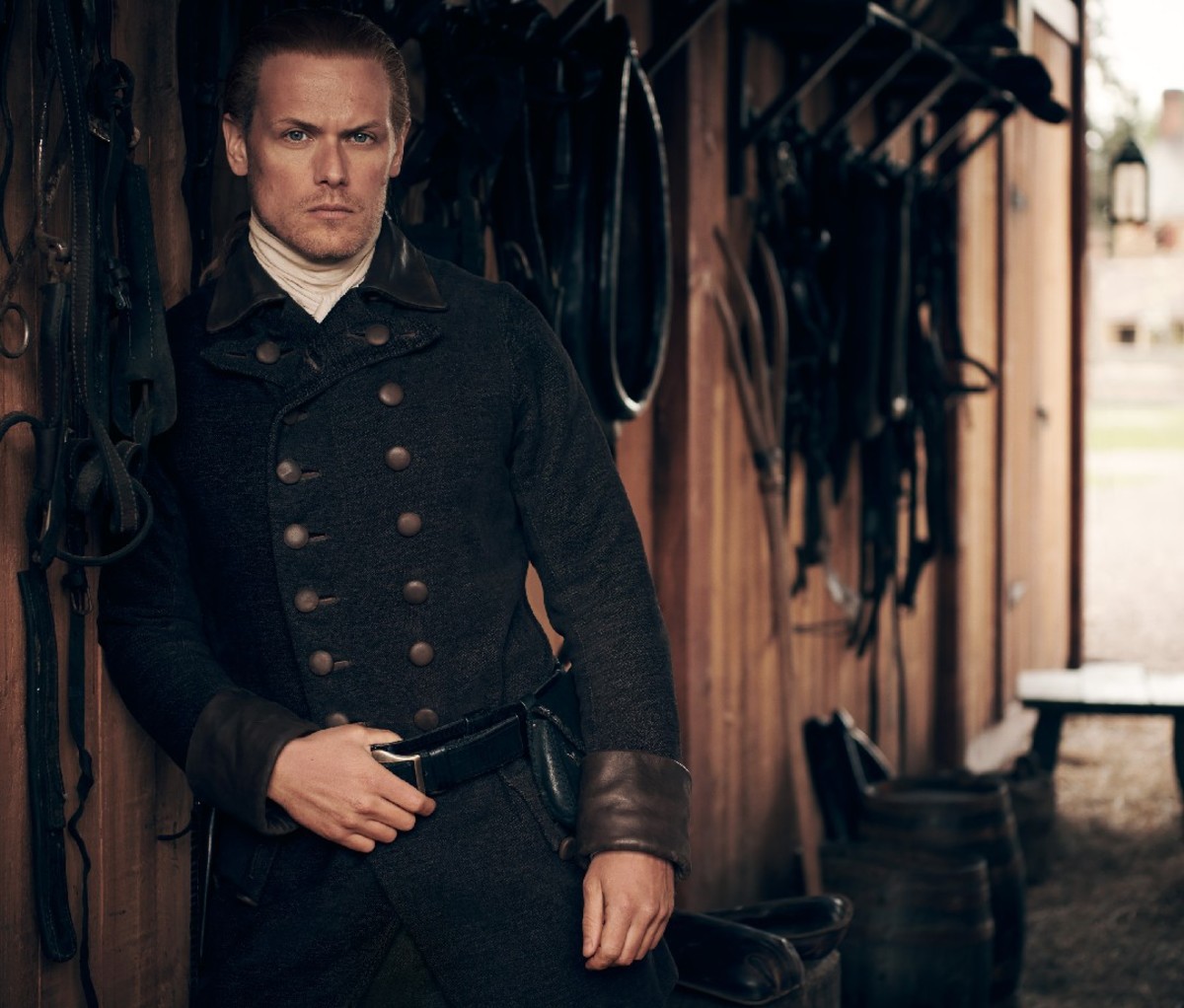 'Outlander' star Sam Heughan stands beside a horse stable wall in character in a dark double breasted jacket