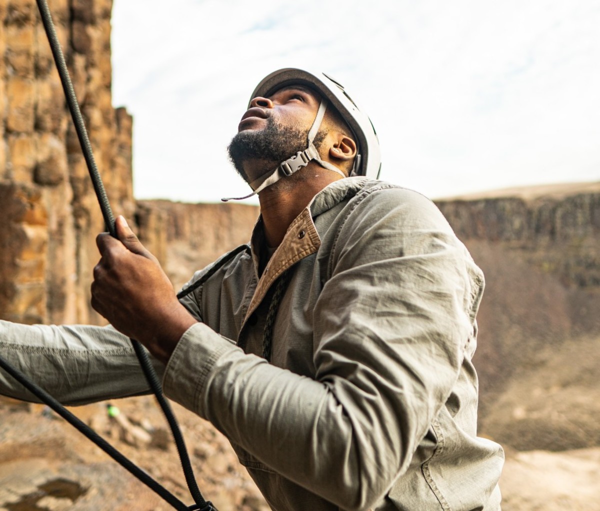 Black man wearing helmet and green jacket holding rope to climb rock wall