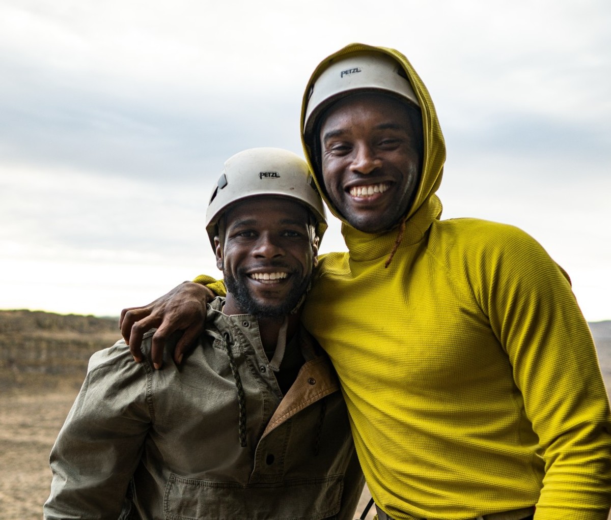 Two Black men wearing climbing helmets smiling at camera with arm around each other