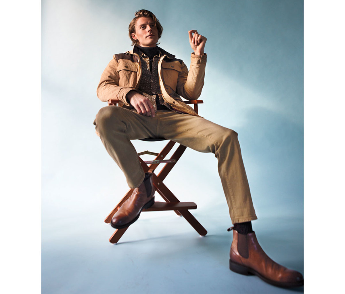 Man sitting in director's chair against blue backdrop wearing tan jacket, pants, and boots