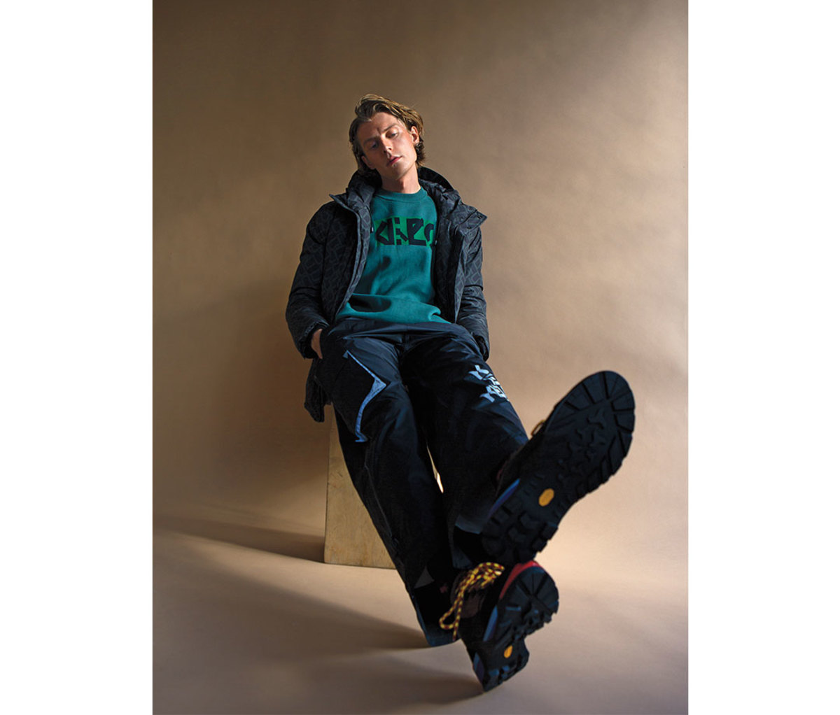 Man leaning against wall wearing hiking boots and blue jacket