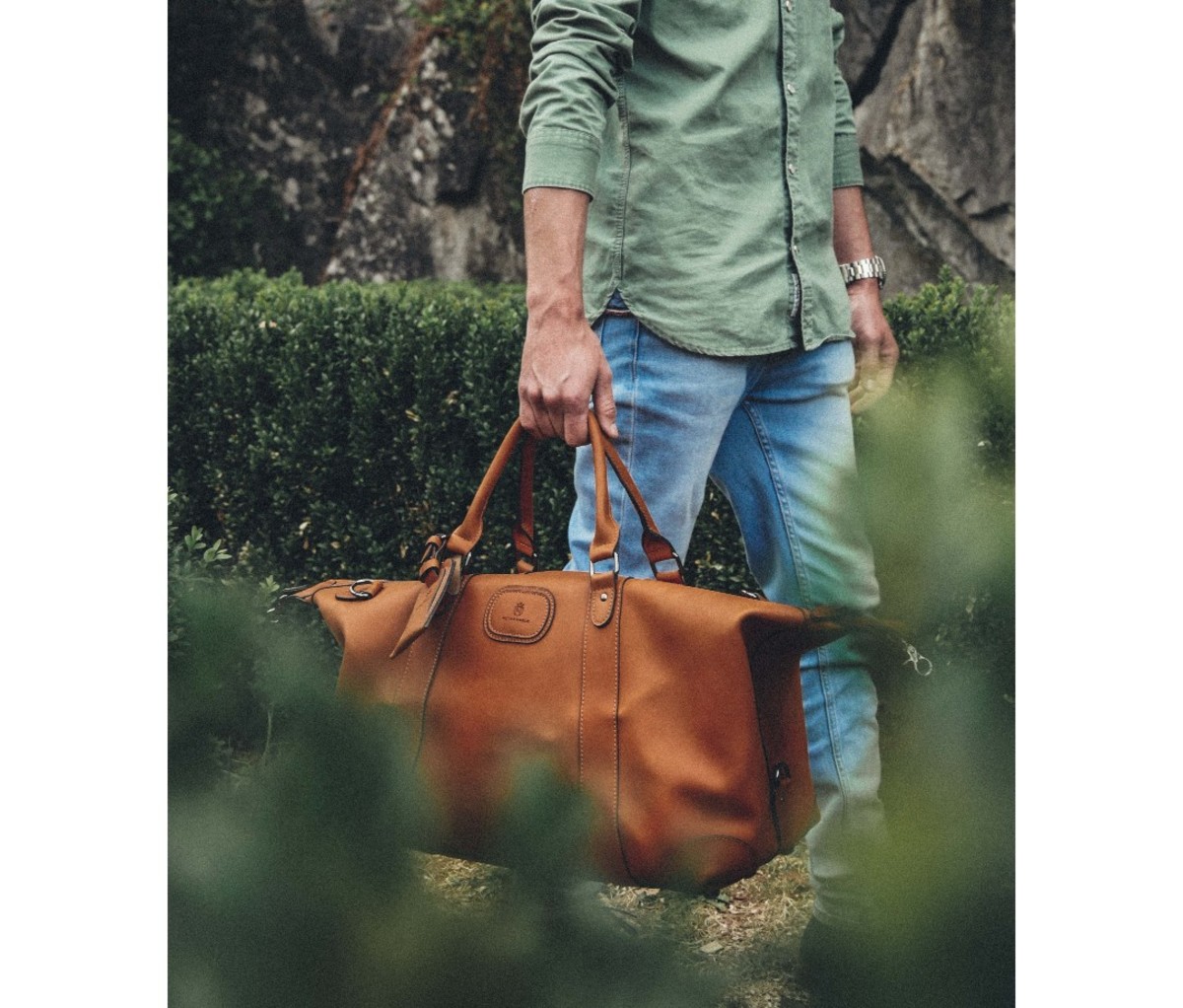 Man in green button-down and jeans holding tan leather duffel bag