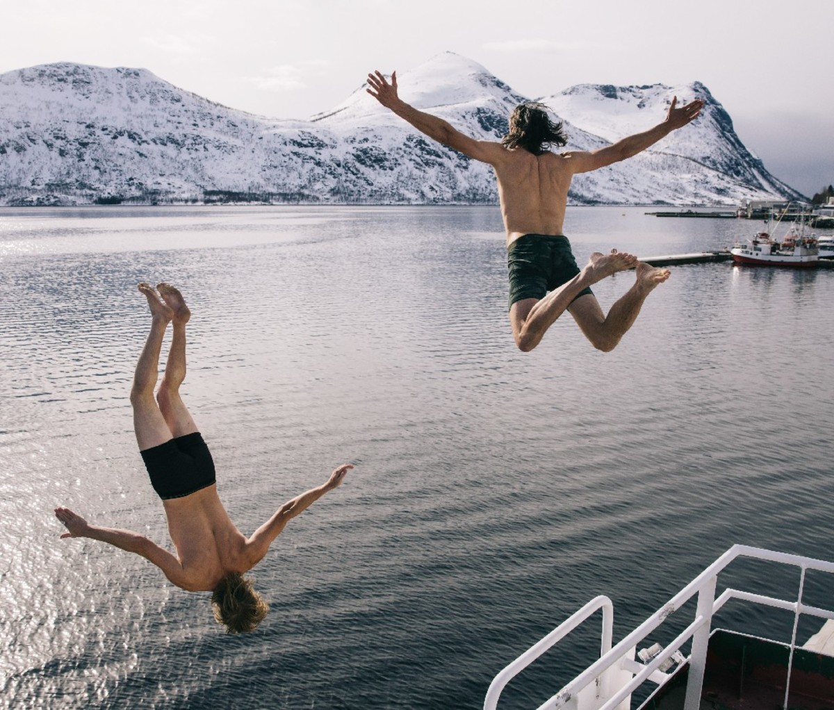 Two guys leaping into the coastal waters of Northern Norway from the vessel, with snowy hillsides in the background.