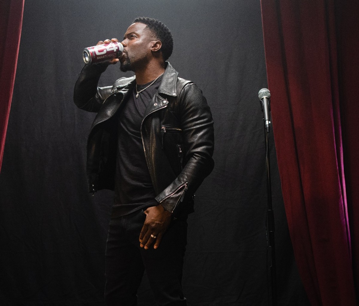 Kevin Hart in a black leather jacket stands next to a mic while drinking a can of C4 energy beverage
