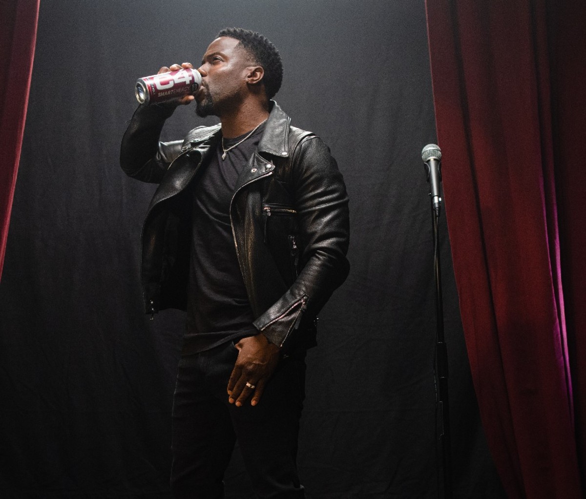 Kevin Hart in a black leather jacket stands next to a microphone while drinking a can of C4 Energy Drink