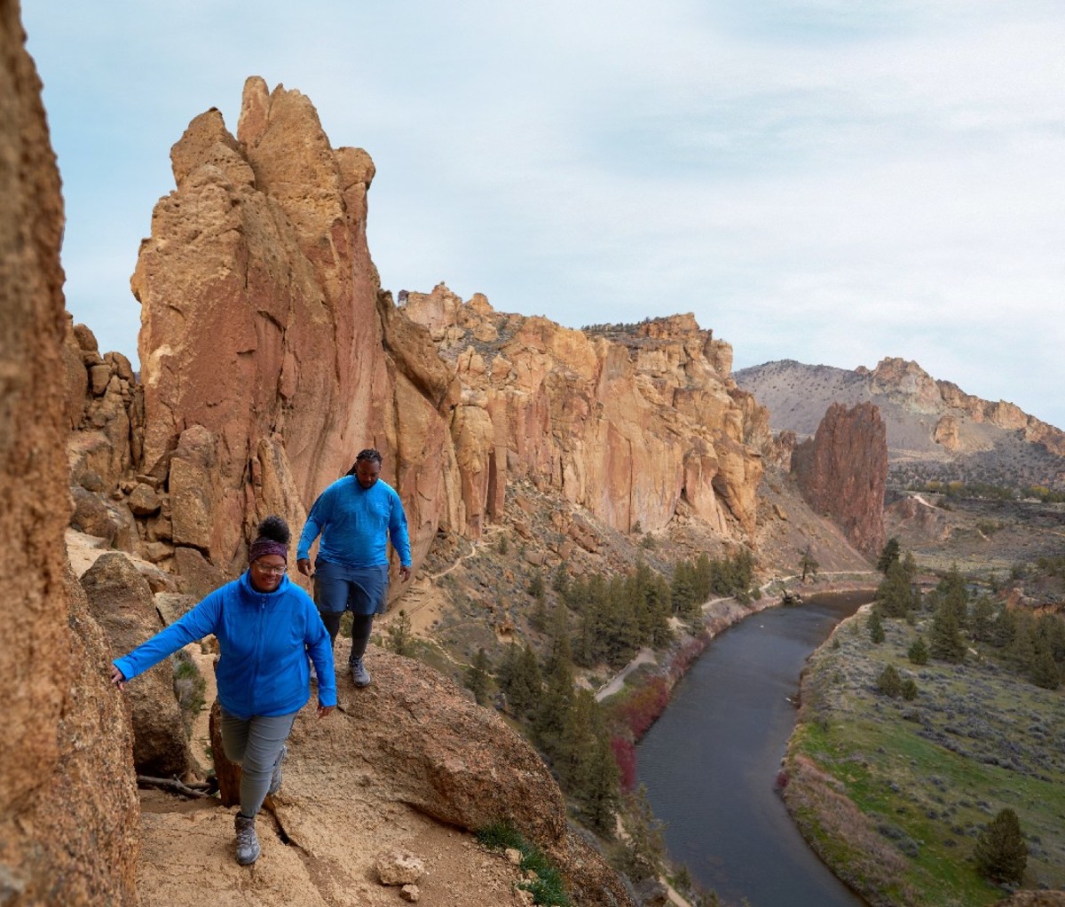 Two hikers in blue jackets walk along a riverside canyon trail.
