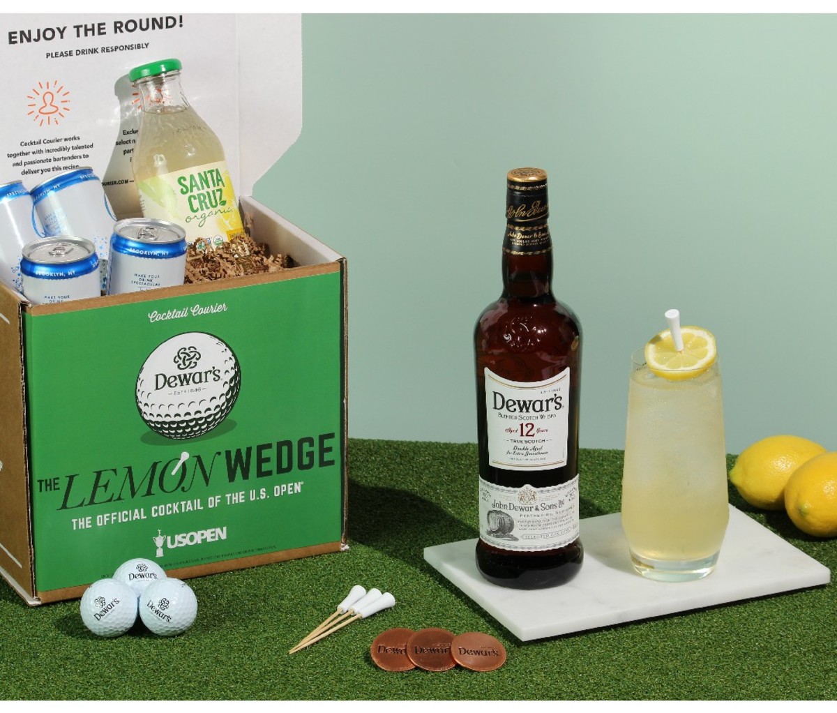Lemon Wedge Cocktail Kit on a table, including a boxed supply of ingredients beside a prepared Lemon Wedge cocktail and a bottle of Dewar's whisky.