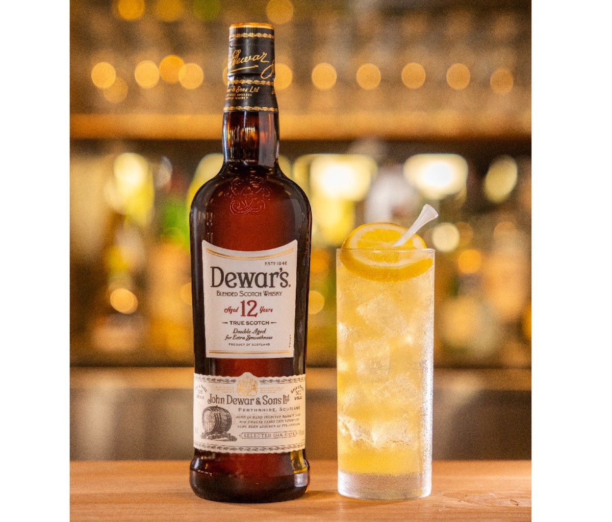 Prepared Lemon Wedge cocktail in a highball glass sits on the bar beside a bottle of Dewar's whisky
