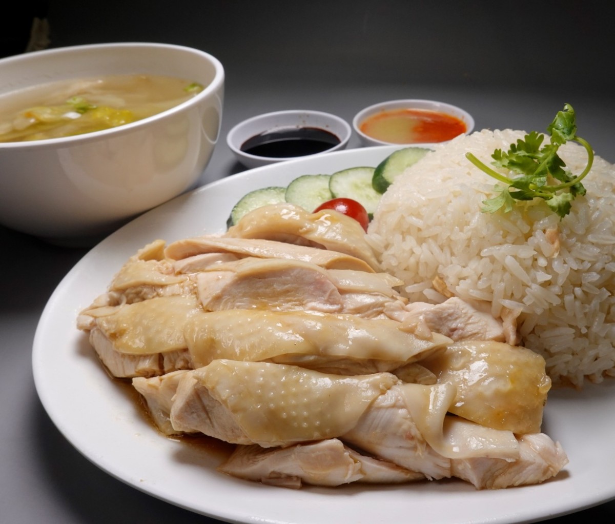 Cut poached chicken with skin next to a pile of rice with green garnish and bowls of dipping sauces in the background. Urban Hawker