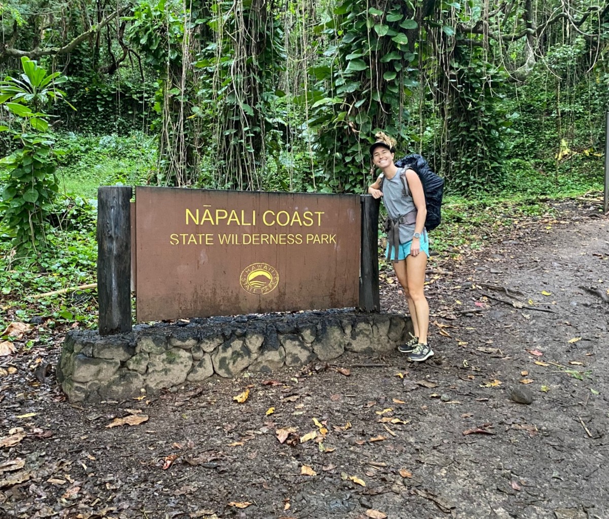 Woman in a backpack and hiking shorts standing next to a Na Pali Coast State Park sign near a muddy path. Thru-hike