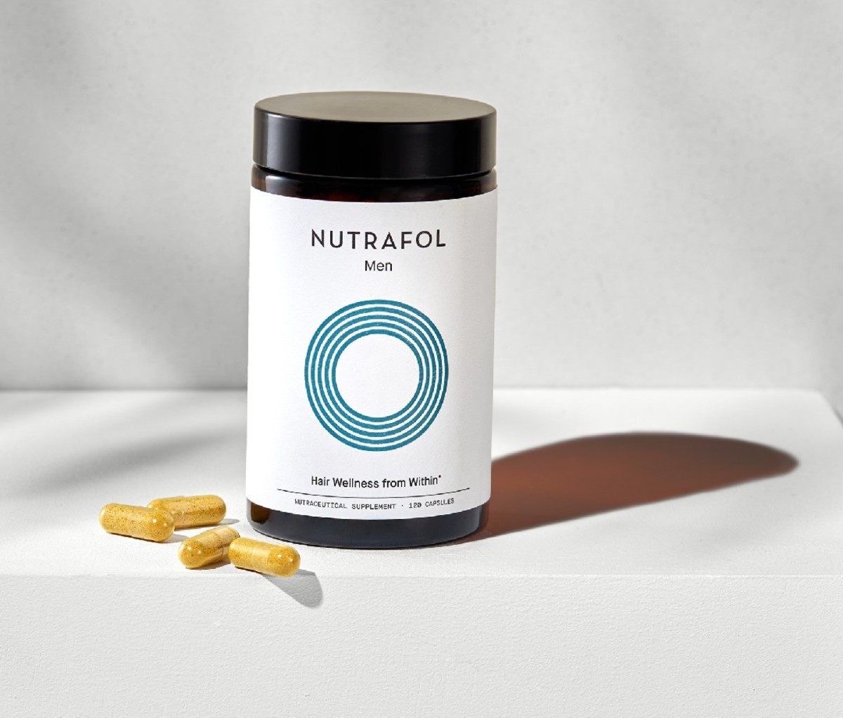 Container of Nutrafol hair loss supplement on a white counter with four loose capsules beside it.