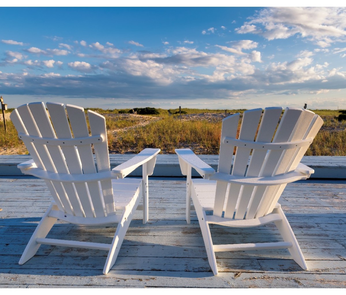 A pair of empty white Adirondack chairs on a porch in Cape Cod overlooking the water.