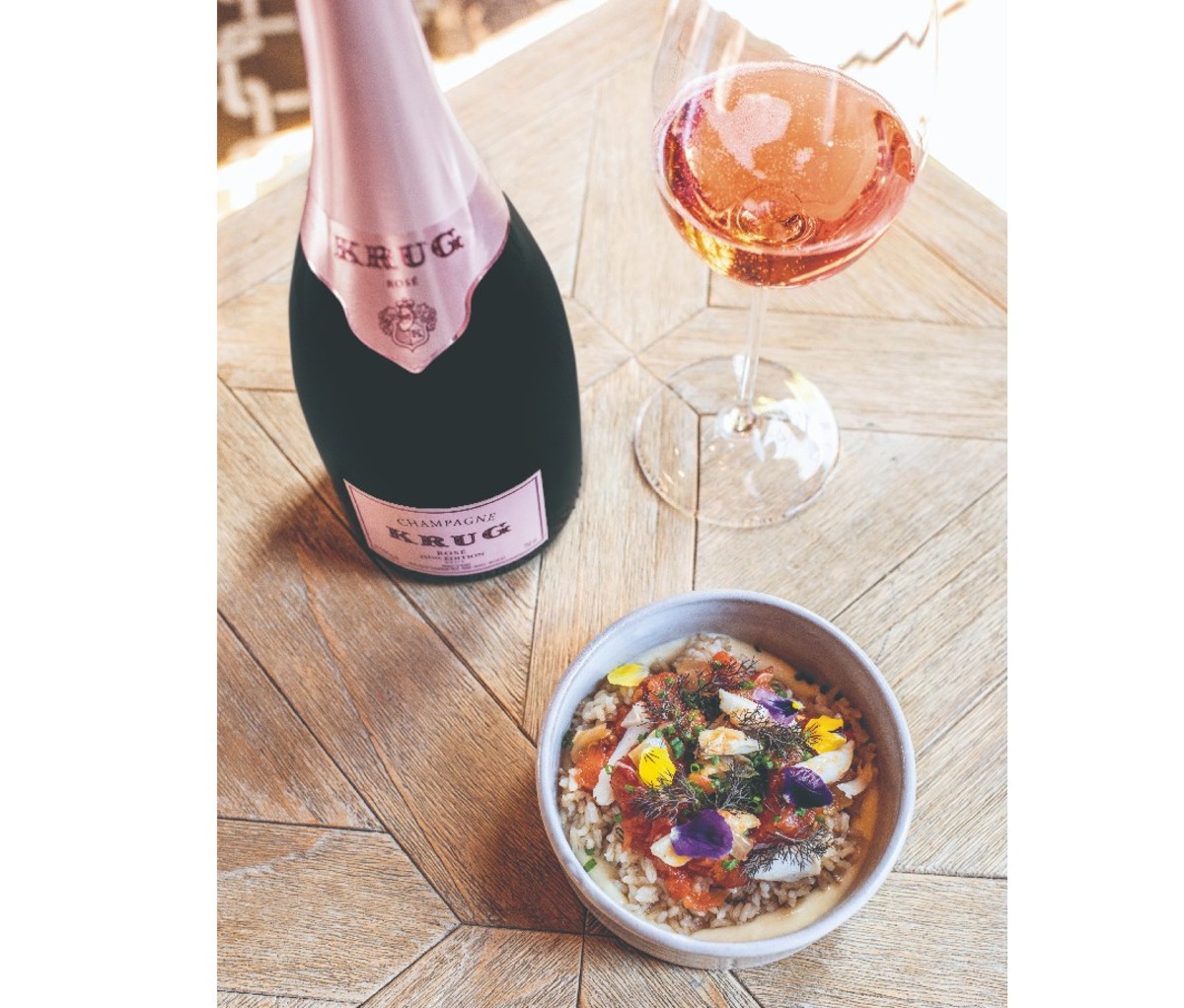 Bottle and filled glass of rosé Champagne on a table beside a paired dish of crispy brown rice with blue crab, smoked tomato vinaigrette and fennel purée