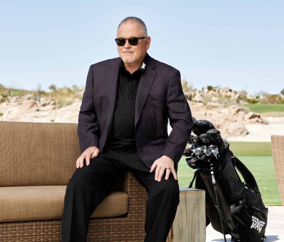 Older caucasian man in. black suit and sunglasses sitting on outdoor sofa next to golf clubs