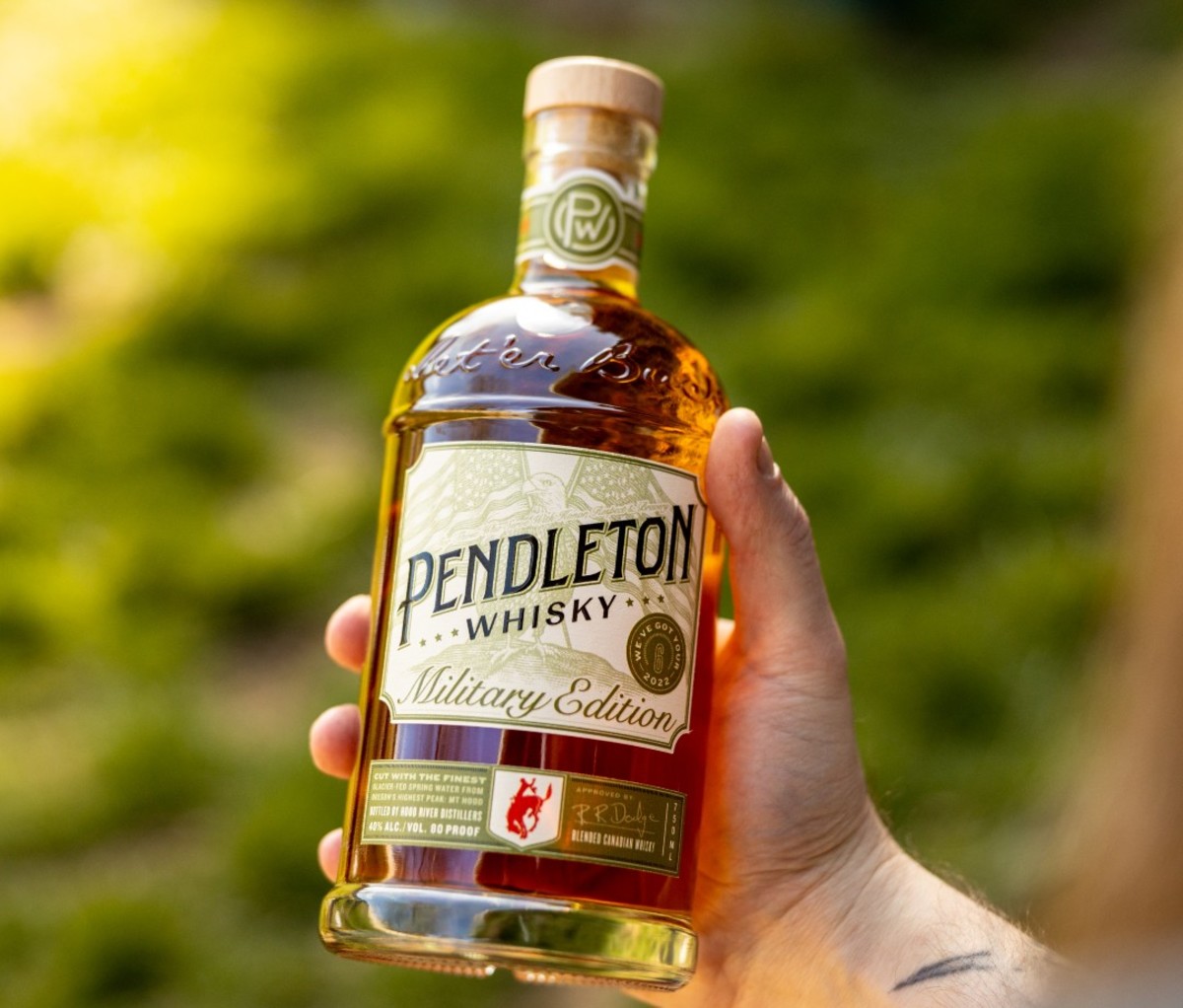 Man's hand holding Pendleton Whiskey bottle outdoors over grass