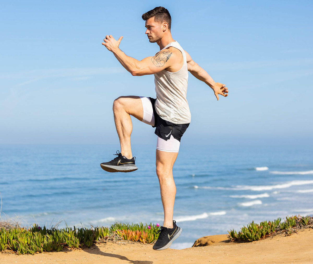 Caucasian man in white tank top and black shorts in jumping position from lunge on beach with ocean in background