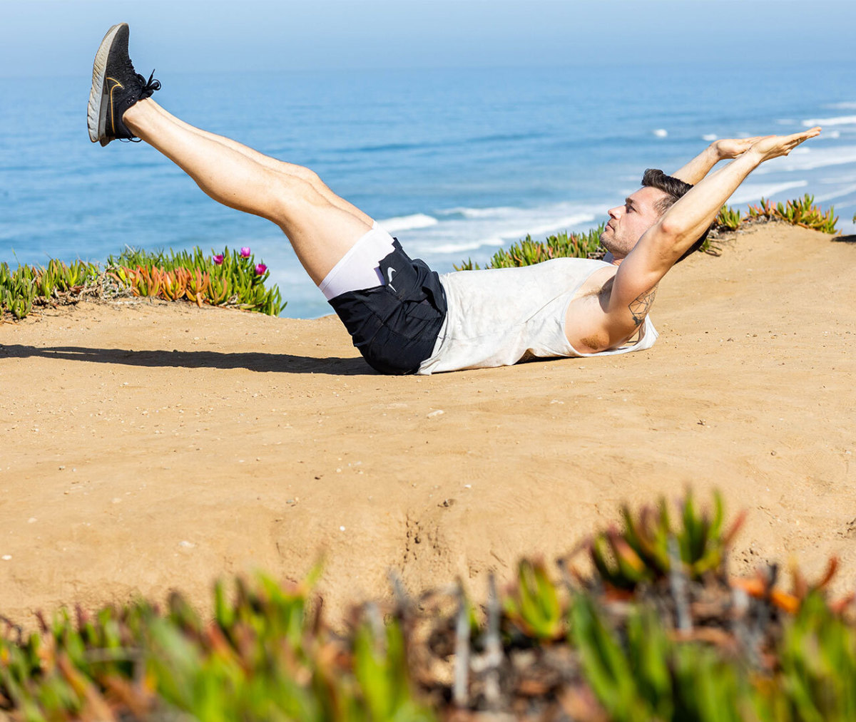 Caucasian man in white tank top and black shorts on back in hollow body hold position with ocean in background