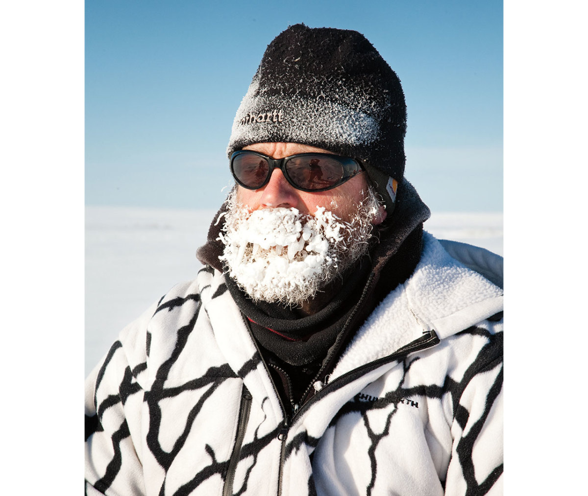 Older caucasian man wearin black banie and sunglasses with white and black fleece and beard covered in ice