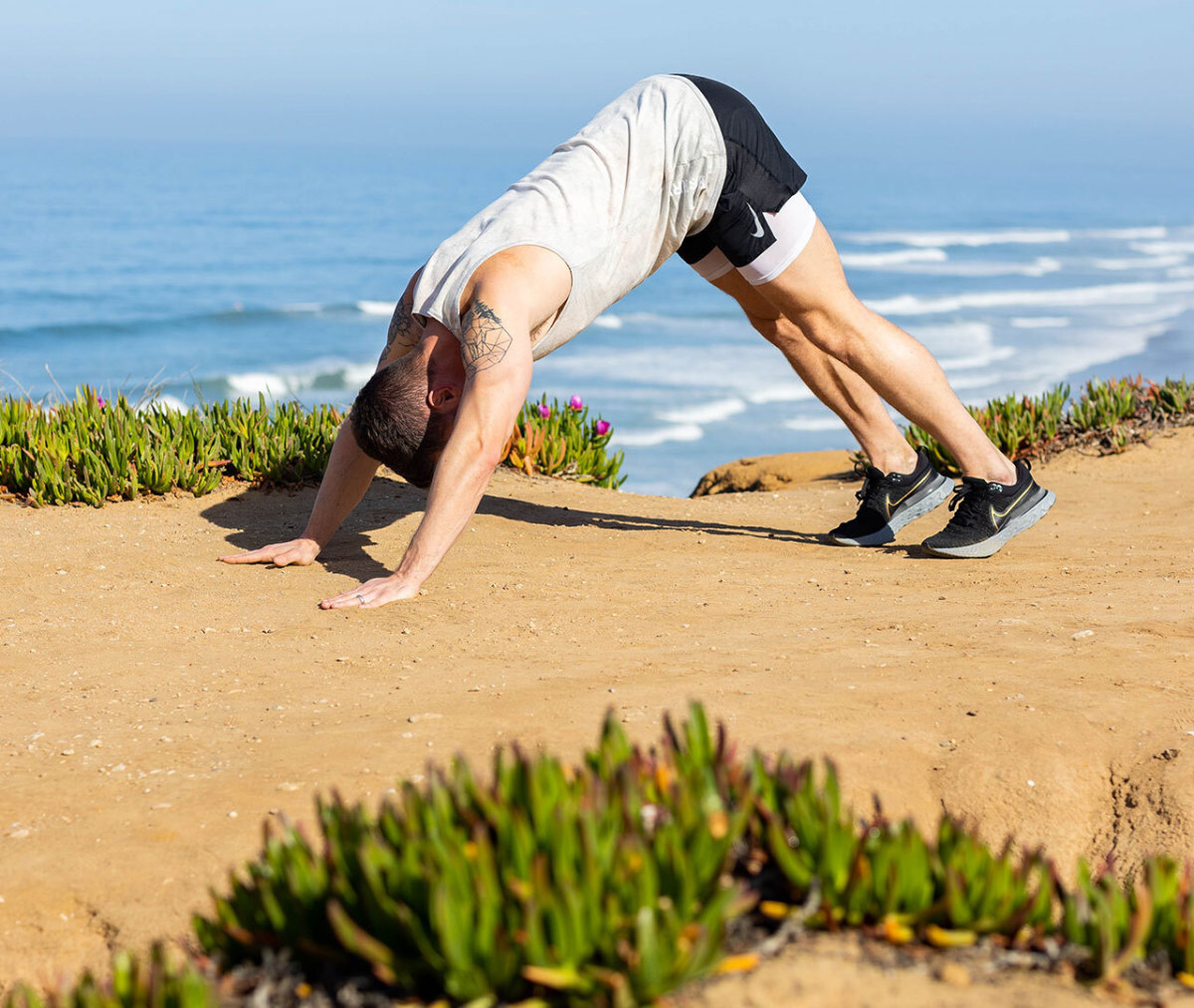 Caucasian man in white tank top and black shorts in pike position on beach with ocean in background