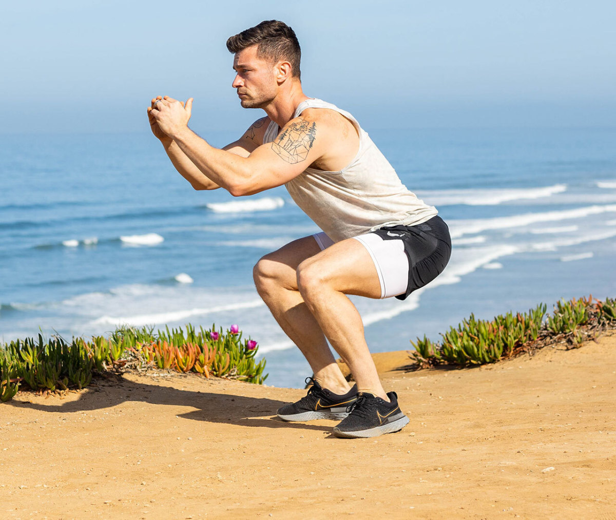 Caucasian man in white tank top and black shorts in squat position on beach with ocean in background