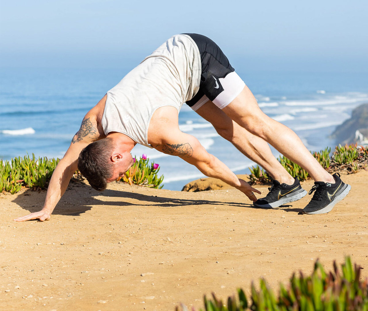 Caucasian man in white tank top and black shorts touching left hand to right foot on beach with ocean in background