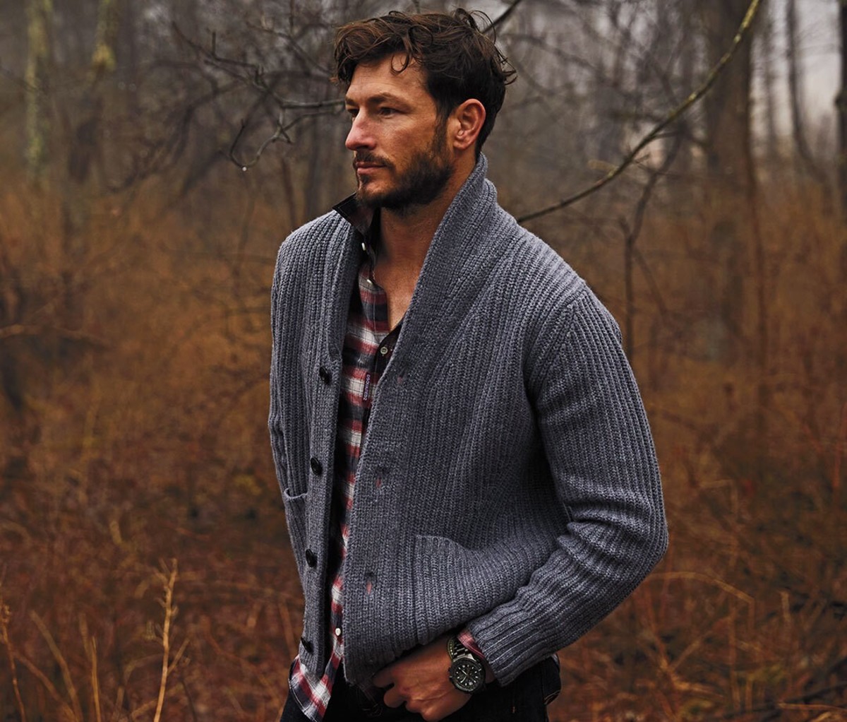 A man in the woods wearing a shirt and cardigan sweater
