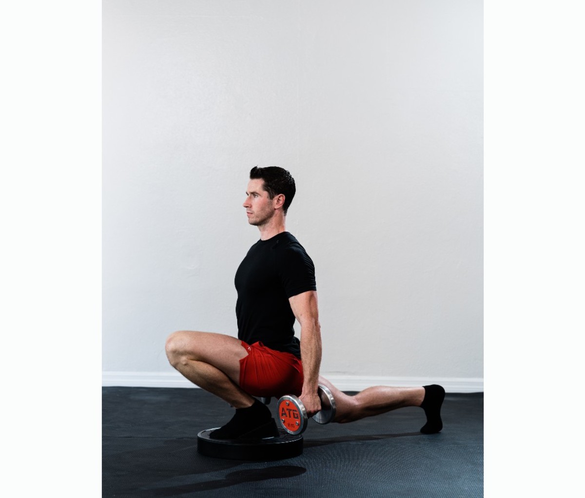 Caucasian man in black T-shirt and red shorts doing exaggerated split squat with dumbbells