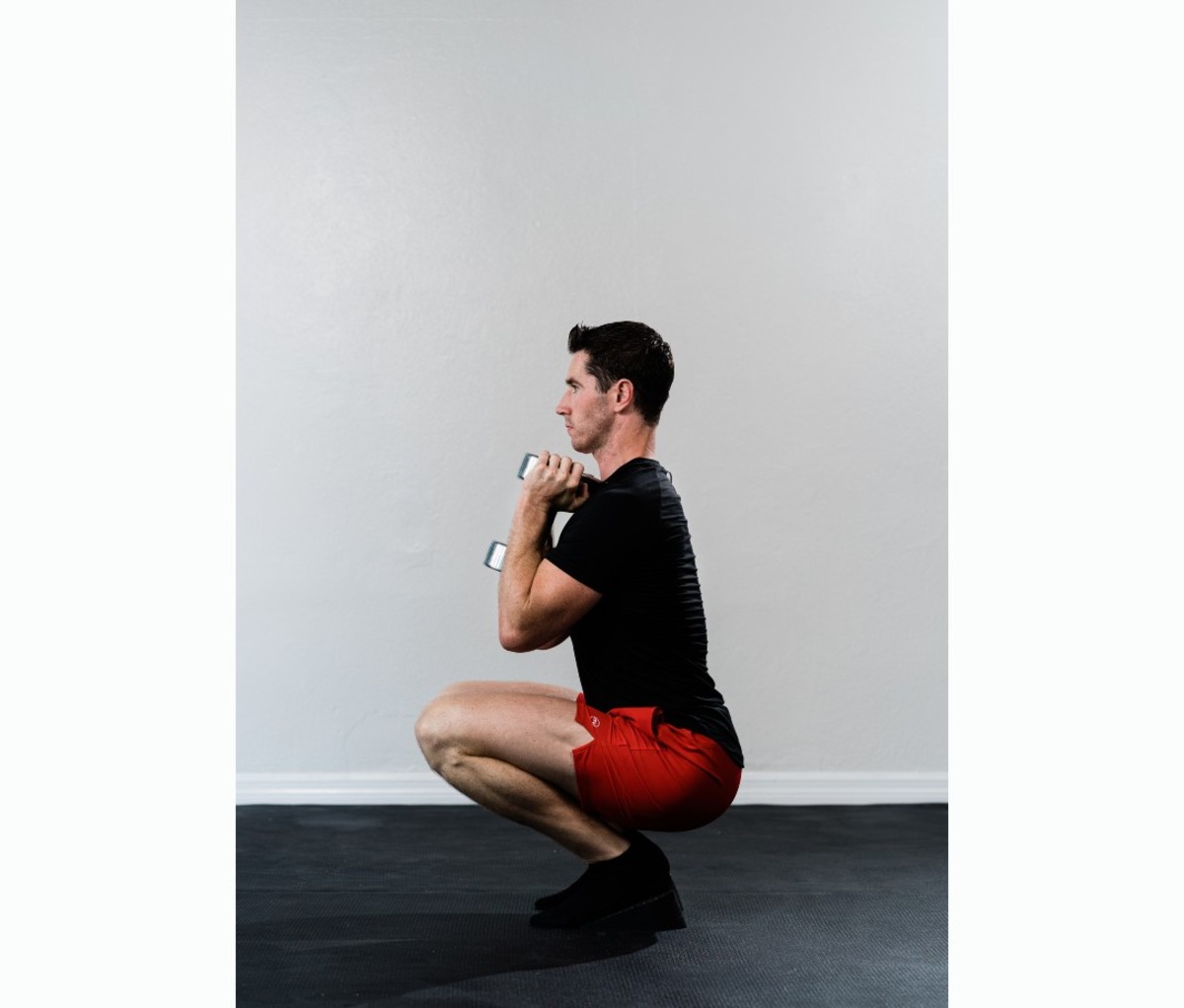 Caucasian man in black T-shirt and red shorts doing heel-elevated squat with dumbbells