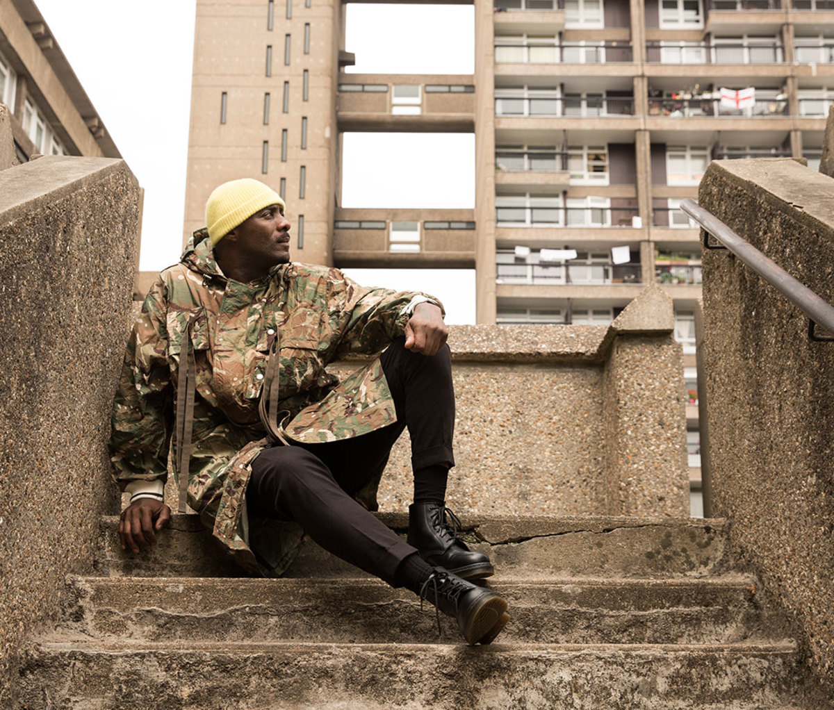 Actor Idris Elba wearing yellow beanie and camo-printed jacket sitting on concrete steps