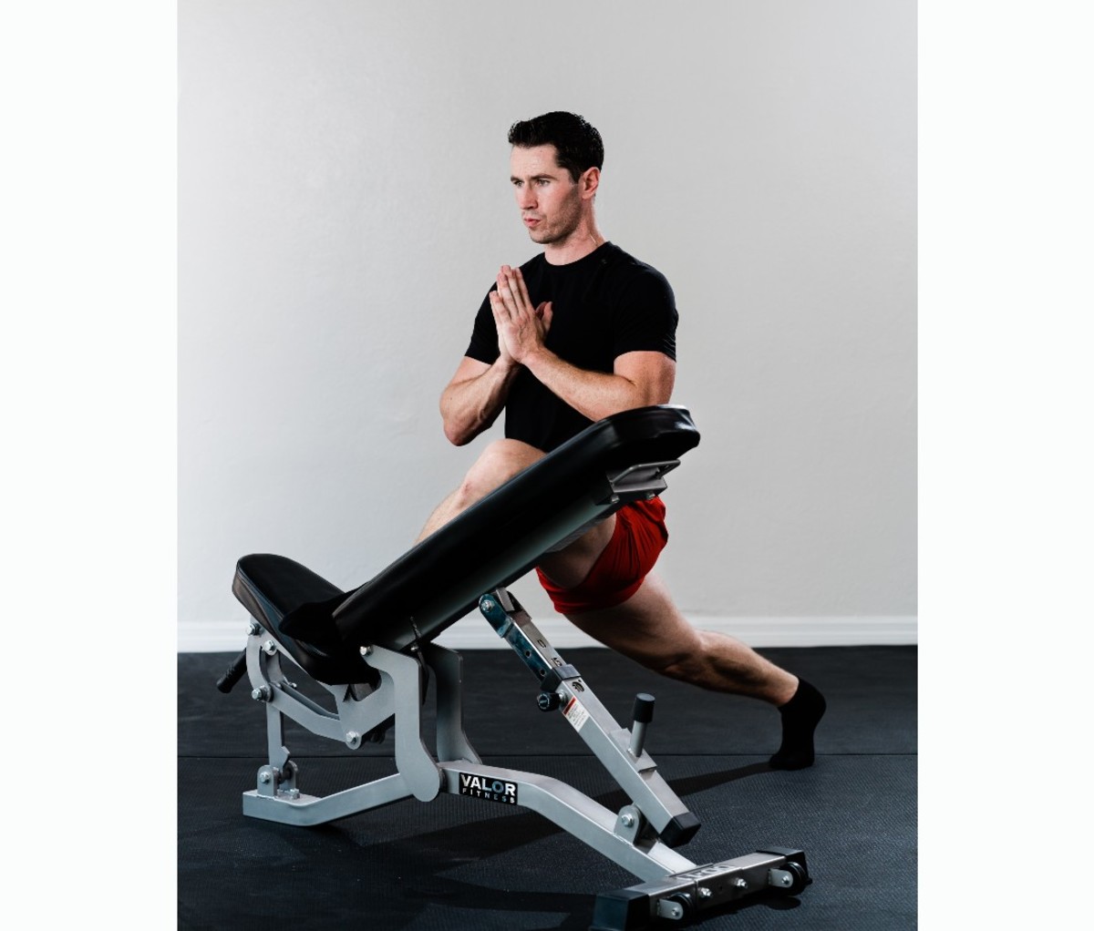 Caucasian man in black t-shirt and red shorts stretching on bench in bent pigeon pose