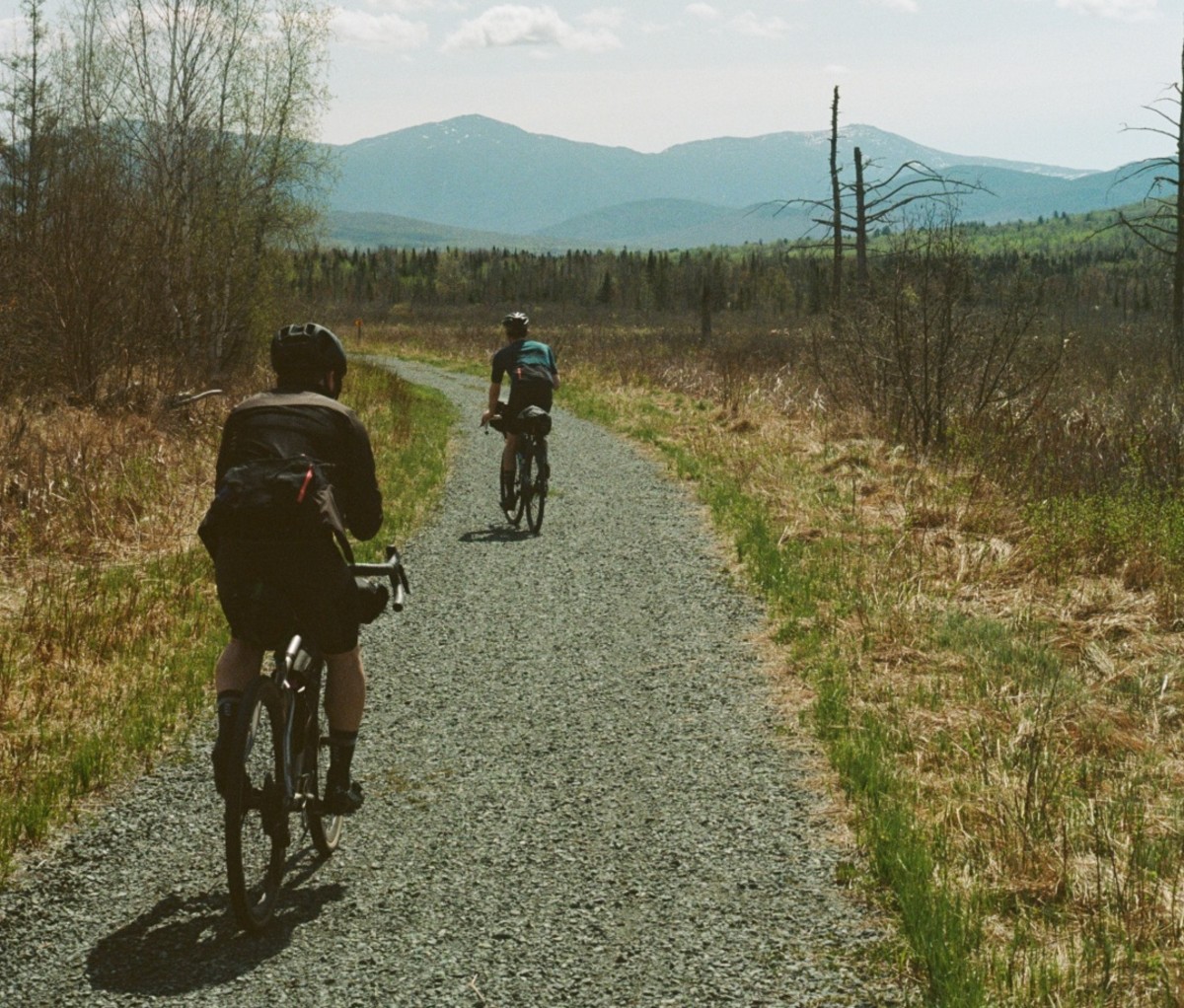 Two cyclists riding on a gravel path with a field and distant mountains in the background. new england bikepacking
