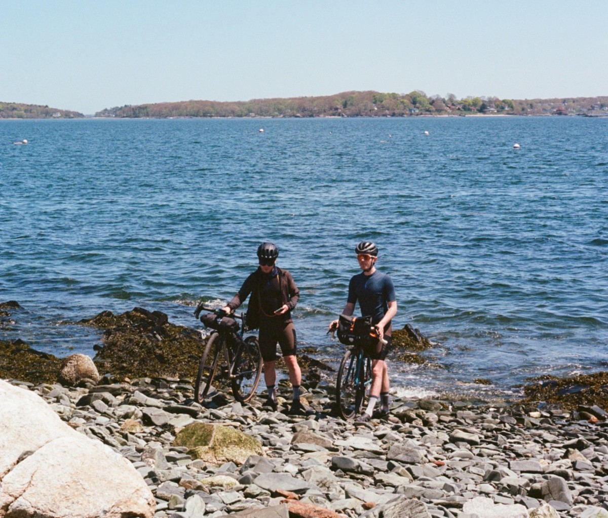 Two cyclists with their bikes standing on a rocky beach with the ocean and land in the background. new england bikepacking