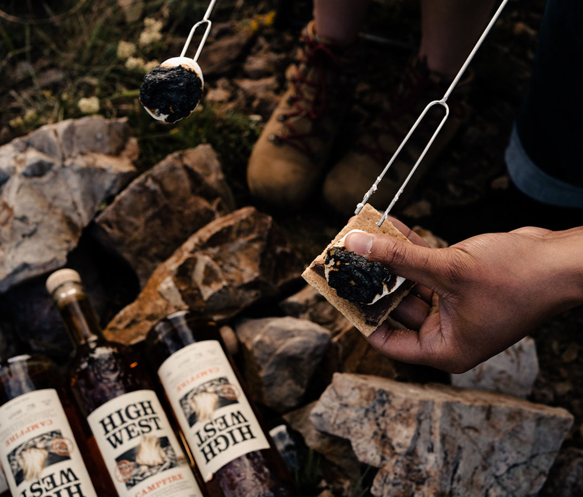 Bottles of High West Campfire whiskey beneath man's hands holding s'mores and stick with marshmallow