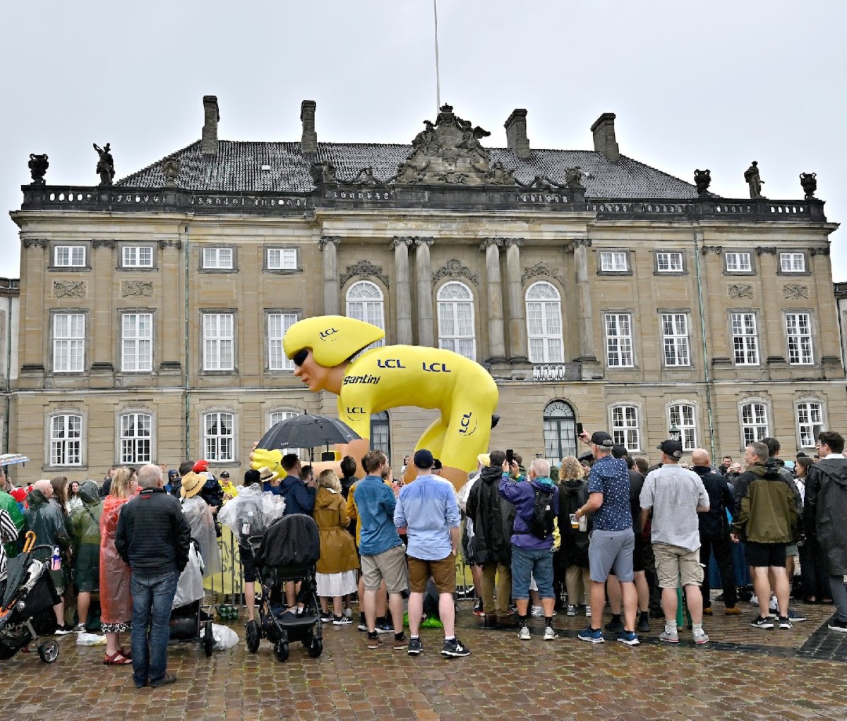 Crowd stands before a historic building in Denmark with a giant yellow-jerseyed blowup cycler