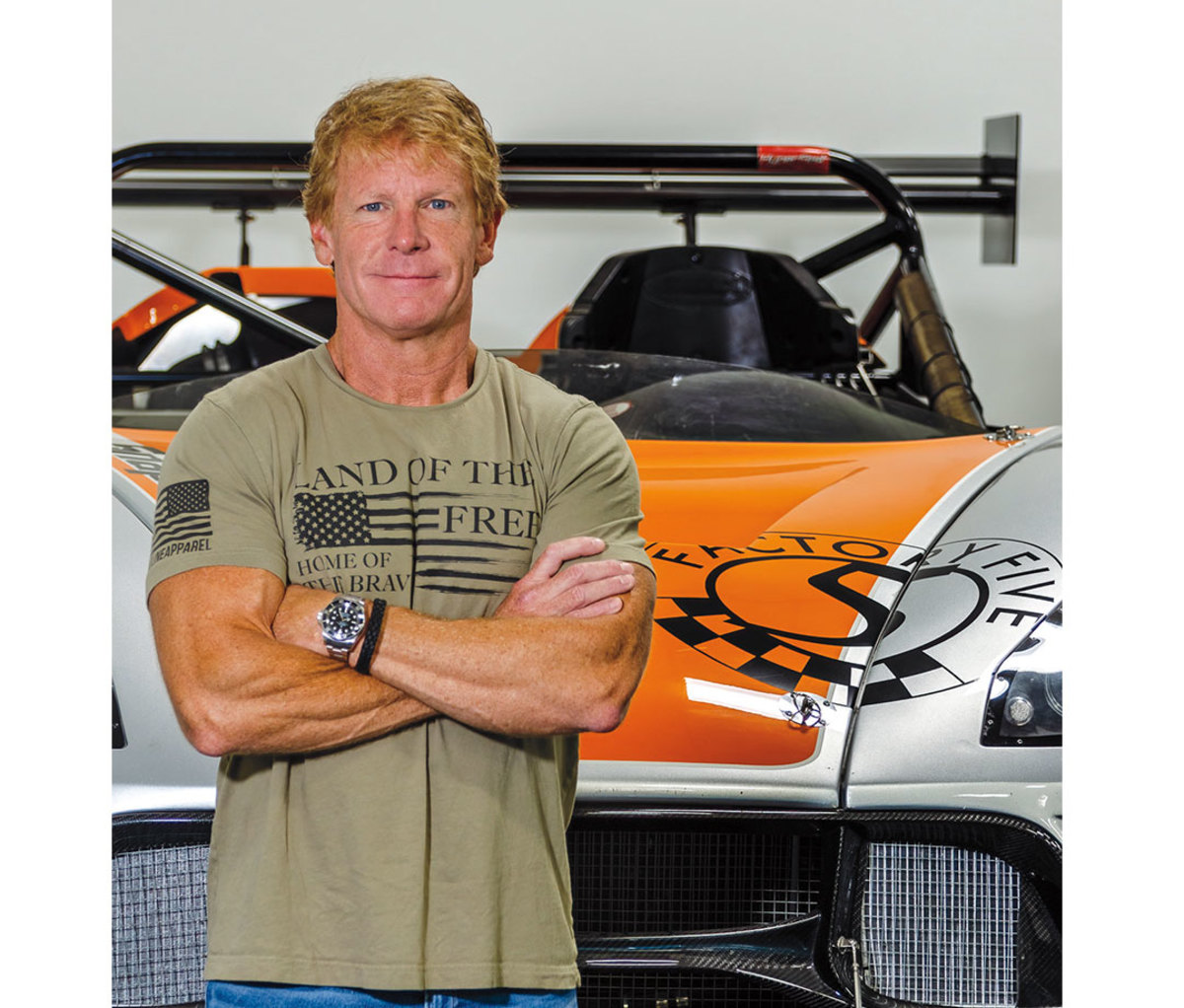 Caucasian man with strawberry blonde hair poses with arms across chest in front of sports car