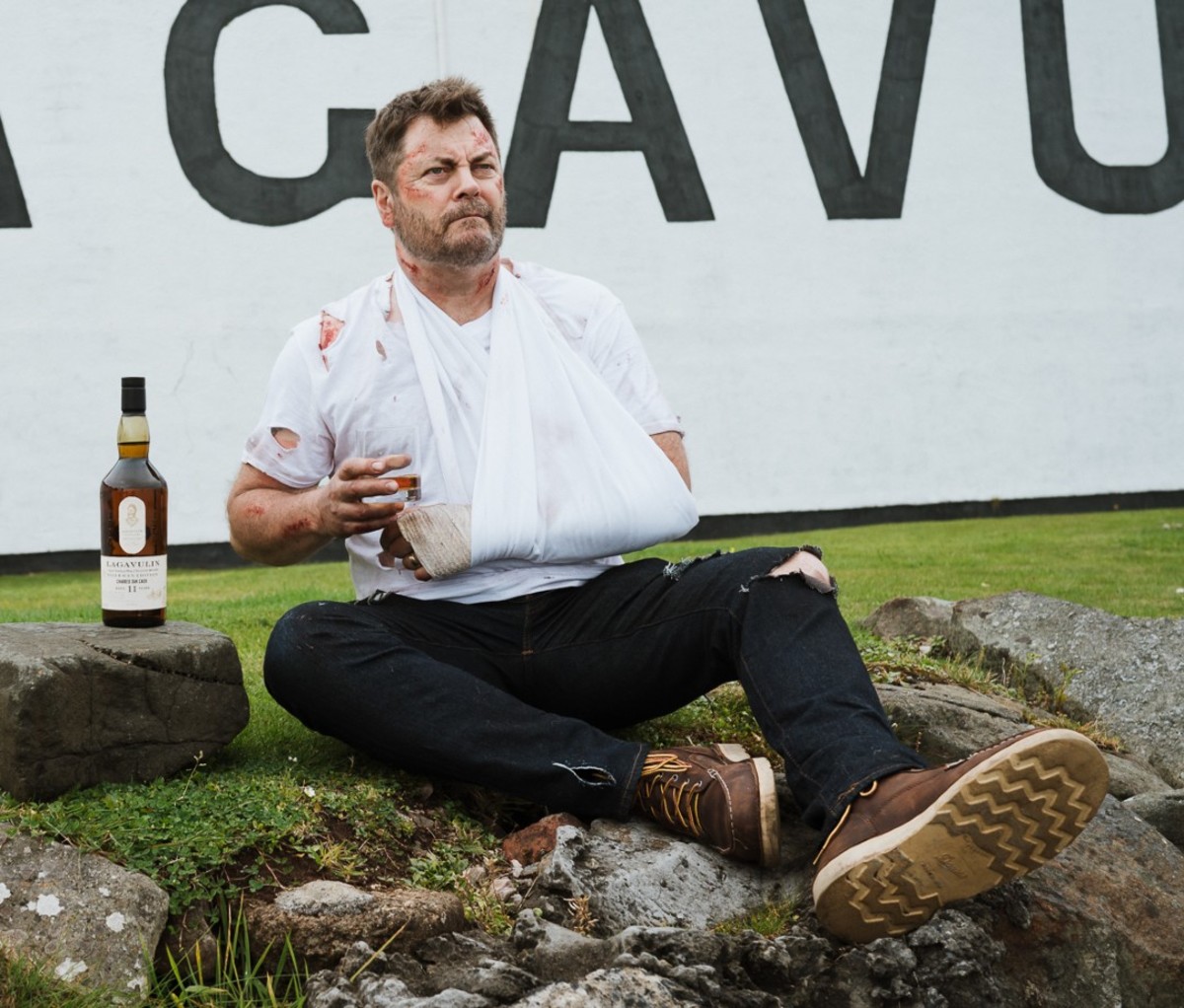Battered man sitting on ground with bottle of Lagavulin Offerman Edition: Charred Oak Cask