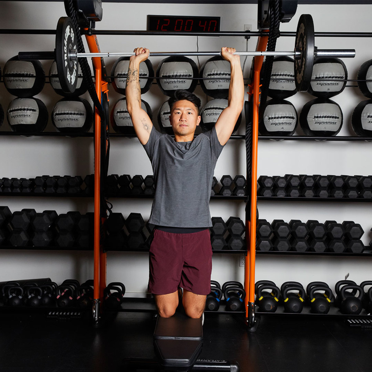 Athletic man wearing gray t-shirt and maroon shorts doing kneeling overhead barbell press