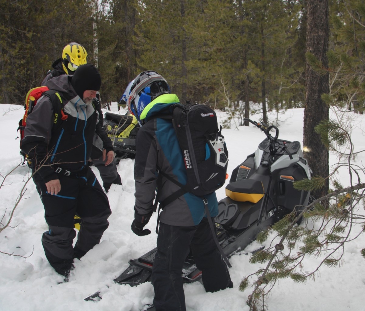 Men standing around a crashed snowmobile next to a tree.