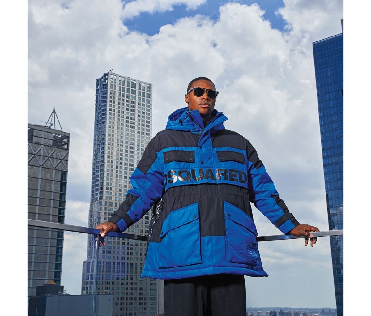 A man wearing a puffer coat stands on a roof in front of a city skyline.