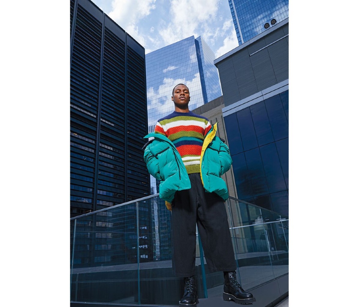 A man wearing a striped sweater and a puffer coat in front of a city skyline.