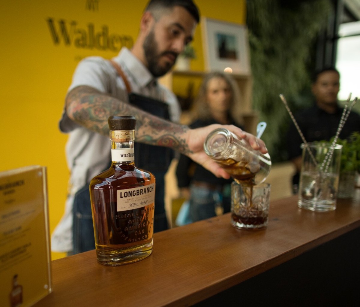 Bartender with tattooed arm pouring cocktail into glass with bottle of Longbranch whiskey on bar