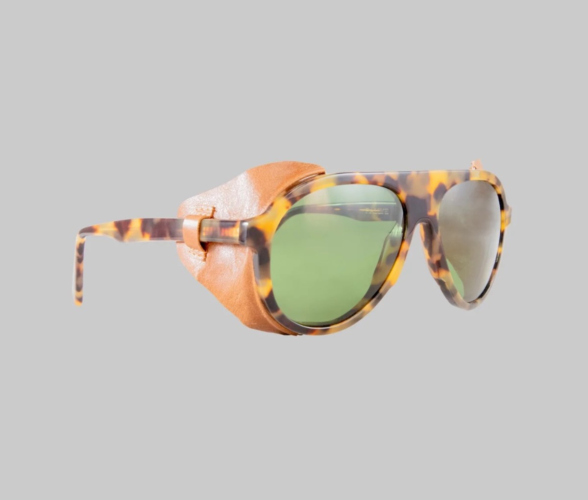 Tortoise shell framed sunglasses with green lenses and leather side hoods on a gray background.