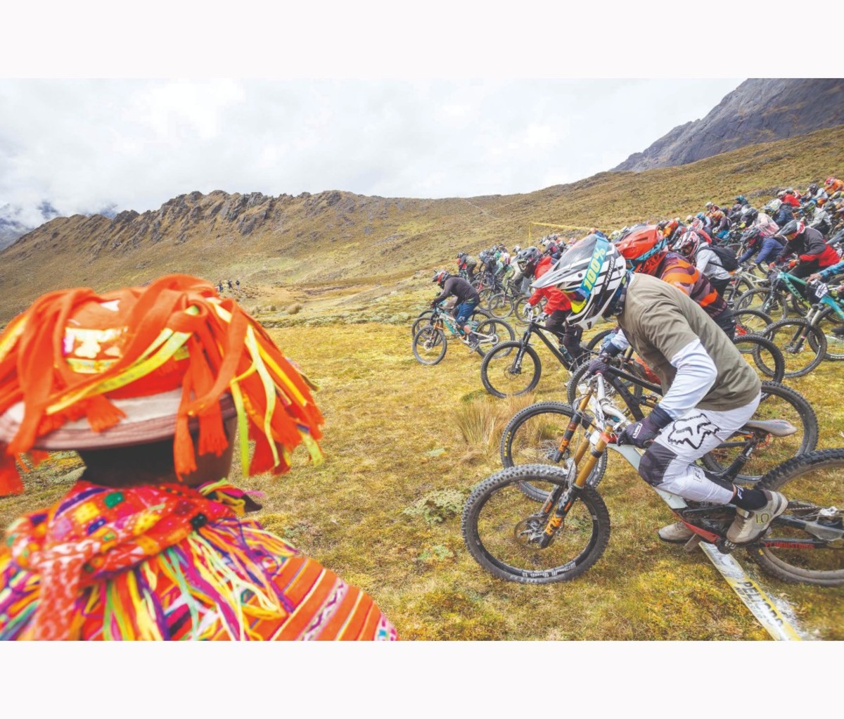 Mountain bikers line up on grass with person in traditional colorful Peruvian garb watches