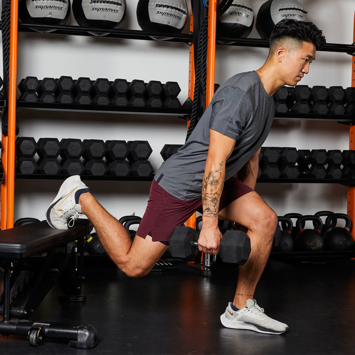 Athletic man wearing gray T-shirt and maroon shorts doing Dumbbell Bulgarian Squat