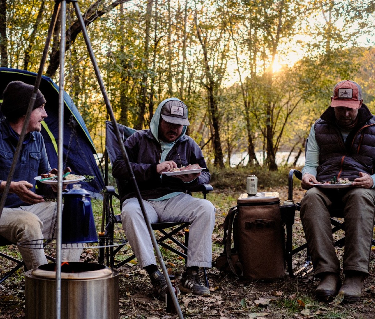 Three guys sitting down to dinner at their campsite in a woodsy river area.