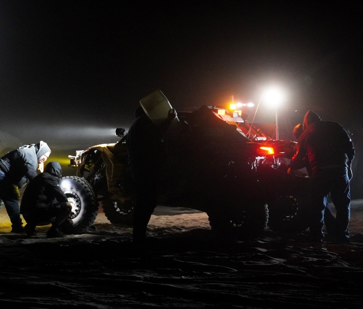 Race car mechanics surround a UTV during a pit stop at night in a desert race.